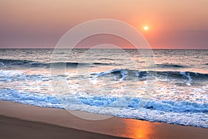 Stunning sunset or sunrise over the sea or ocean on the beach, purple sky, blue waves, white foam and golden sun reflection