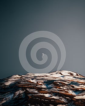 Stunning view of a daytime half-moon on the blue sky above the snow-capped mountain during sunset