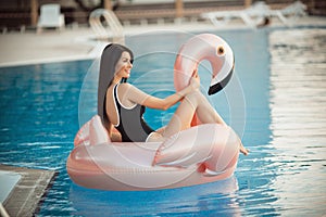 Stunning woman is wearing black bikini sitting in swimming pool with blue water on a pink flamingo mattress, summer