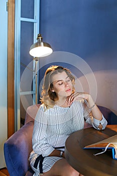 Stunning young girl looks away with downcast eyes, holds hand near face and sits in chair at table on background of lamp