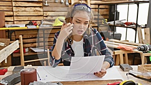 Stunning young hispanic female carpenter absorbed in her work, sending a voice message via smartphone while reading construction