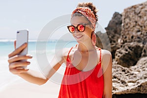 Stunning young lady in red attire using phone for selfie at wild beach. Adorable white girl in spar