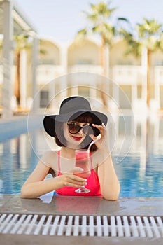 Stunning young lady in trendy pink swimsuit, sitting on the edge of pool and enjoying cocktail