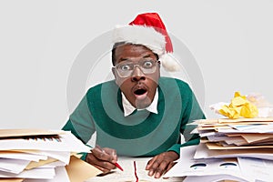 Stupefied dark skinned male opens mouth with amazement, wears spectacles and red festive hat, surprised with many tasks