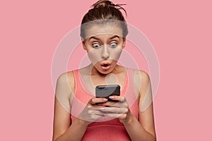 Stupefied emotional woman stares with bugged eyes at smart phone, feels shocked as reads news at web page, connected to