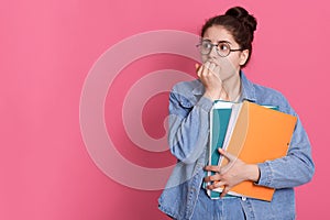 Stupefied frightened schoolgirl holds colorful folders with papers, being shocked, dressed in casual denim jacket, poses 