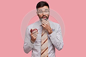 Stupefied teacher with thick bristle holds chin, has snack between classes, dressed in formal shirt with tie, bites