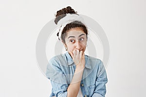 Stupefied woman with dark in do-rag and denim shirt has upset expression, being confused or worried, bites her