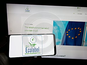 Person holding cellphone with logo of environmental certification EU Ecolabel on screen in front of webpage.