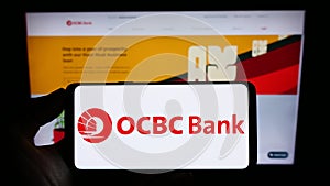 Person holding cellphone with logo of Oversea-Chinese Banking Corporation (OCBC Bank) on screen in front of webpage.