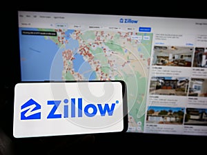 Person holding cellphone with logo of US real estate marketplace Zillow on screen in front of business webpage.