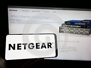 Person holding mobile phone with logo of American computer networking company Netgear Inc. on screen in front of web page.