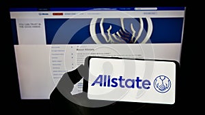 Person holding mobile phone with logo of American insurance company The Allstate Corporation on screen in front of webpage.