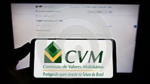 Person holding mobile phone with logo of Brazilian Comissao de Valores Mobiliarios (CVM) on screen in front of web page.