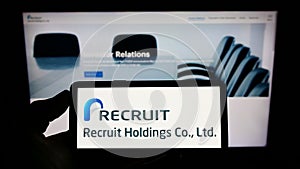 Person holding mobile phone with logo of Japanese company Recruit Holdings Co. Ltd. on screen in front of web page.