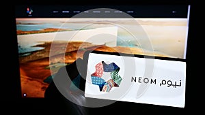 Person holding mobile phone with logo of Saudi Arabian property developer NEOM Company on screen in front of web page.
