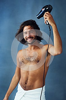 Styling his crowning glory. Studio portrait of a handsome young man blowdrying his hair against a blue background.