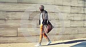 Stylish african man model looking away wearing black leather rock jacket and bag walking on evening city street background