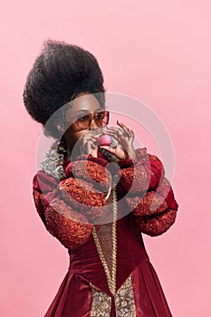 Stylish african woman in image of medieval royal person, wearing red dress and sunglasses, blowing balloon against pink