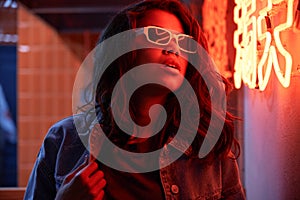 Stylish African woman wears trendy glasses stands near neon sign in night club.