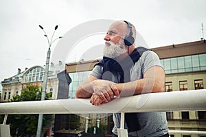 Stylish aged man in headphones leaning on handhold and musing