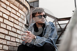 Stylish American young man hipster in trendy blue denim clothes in black sunglasses sits on a vintage staircase near brick wall