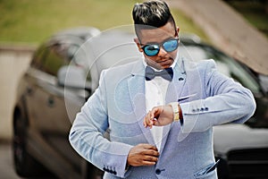 Stylish arabian man in jacket, bow tie and sunglasses against black suv car. Arab rich businessman looking on watches