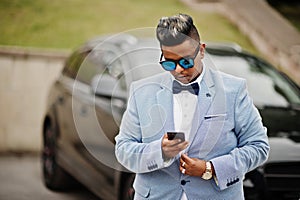 Stylish arabian man in jacket, bow tie and sunglasses against black suv car. Arab rich businessman with mobile phone