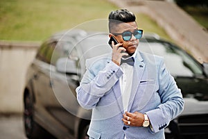 Stylish arabian man in jacket, bow tie and sunglasses against black suv car. Arab rich businessman speaking on mobile phone