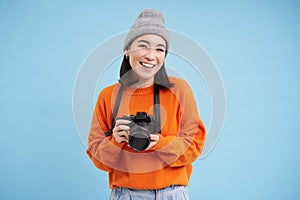 Stylish asian girl with digital camera, taking pictures. Woman photographer smiling, standing over blue background