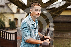 Stylish attractive man in a fashionable denim jacket in a white t-shirt is standing and smiling near a vintage wrought fence