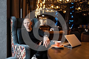 Stylish attractive young man in a fashionable shirt in stylish black jeans in sneakers sits on an easy chair in a restaurant