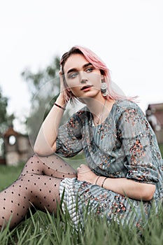 Stylish attractive young woman with pink hair in trendy blue floral dress is relaxing on green grass outdoors on summer day. Cute