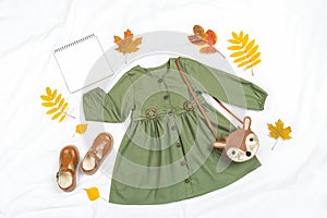 Stylish autumn set of child clothes. Green dress, brown bag, shoes, blank notebook mockup and autumn leaves on white background.