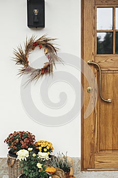 Stylish autumn wreath hanging at modern lantern, pumpkins and flowers at wooden door outdoor. Fall decor and arrangement of