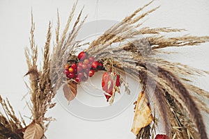 Stylish autumn wreath hanging on white wall outdoor. Close up of rustic autumn wreath with dried grass, berries, herbs and leaves