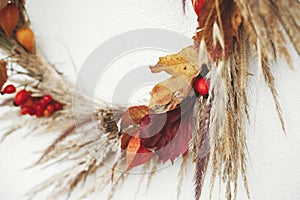 Stylish autumn wreath hanging on white wall outdoor. Close up of rustic autumn wreath with dried grass, berries, herbs and leaves