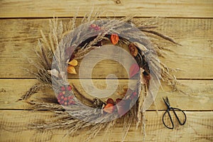 Stylish autumn wreath on rustic table flat lay. Rustic autumn wreath with dried grass, berries, herbs and scissors on wooden table