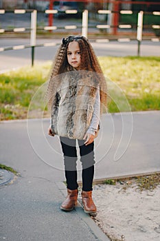 Stylish baby girl 4-5 year old wearing boots, fur coat standing in park. Looking at camera. Autumn fall season