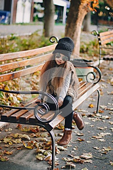 Stylish baby girl 4-5 year old wearing knitted hat, sunglasses, boots, fur coat sitting on the bench in park. Looking at camera.