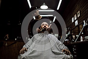 The stylish barbershop. The fashion barber tidies up beard of brutal man sitting in the armchair
