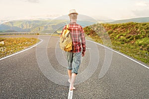 Stylish barefoot bearded male hitchhiker traveler in a hat and with a backpack walks along a country road in the