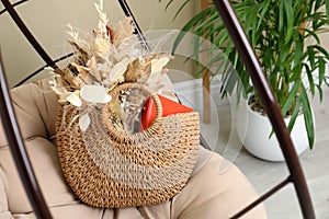 Stylish beach bag with beautiful bouquet of dried flowers and book on papasan chair indoors