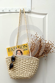 Stylish beach bag with beautiful dried flowers, sunglasses and magazine hanging on white wooden door
