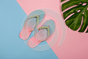 Stylish beach flip-flops on pink and blue pastel background with monstera leaf, top view. Summer concept with copy space