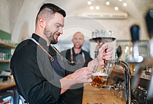 Stylish bearded barman dressed black uniform beer tapping at bar counter and waiter with tray chatting smiling to each other.