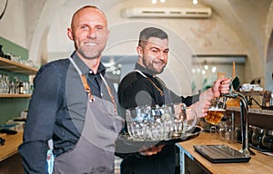 Stylish bearded barman dressed black uniform beer tapping at bar counter and waiter with tray chatting smiling to each other.