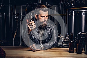 Stylish bearded biker dressed black leather jacket sitting at bar counter in indie brewery.