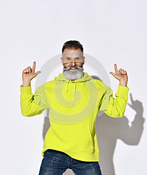 Stylish bearded middle aged man hipster in yellow hoodie and glasses dances with fingers up, points up at copy space