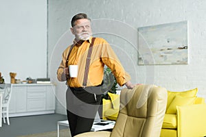 stylish bearded senior man holding cup of coffee and looking away while standing near armchair